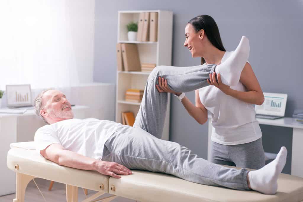 female practitioner in white shirt helping male patient lift left leg up while lying down on bed